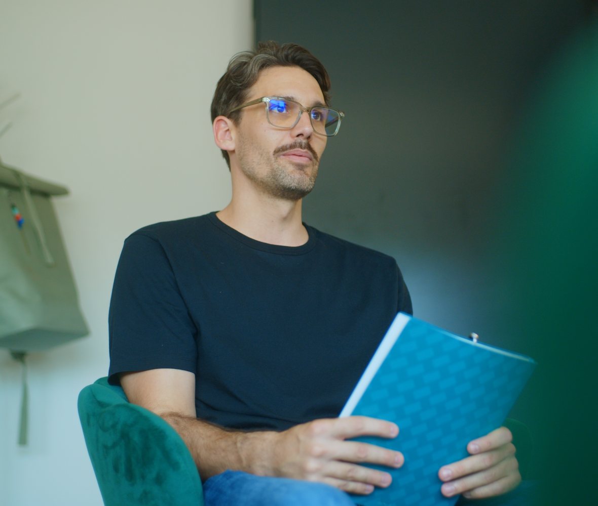 Roger Gloor sitzt mit einem blauen Ordner im Sessel und hört zu